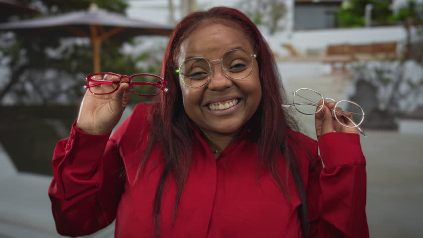 Woman wearing glasses smiles while holding red and clear eyeglass frames near face on street; fashion decision joy.