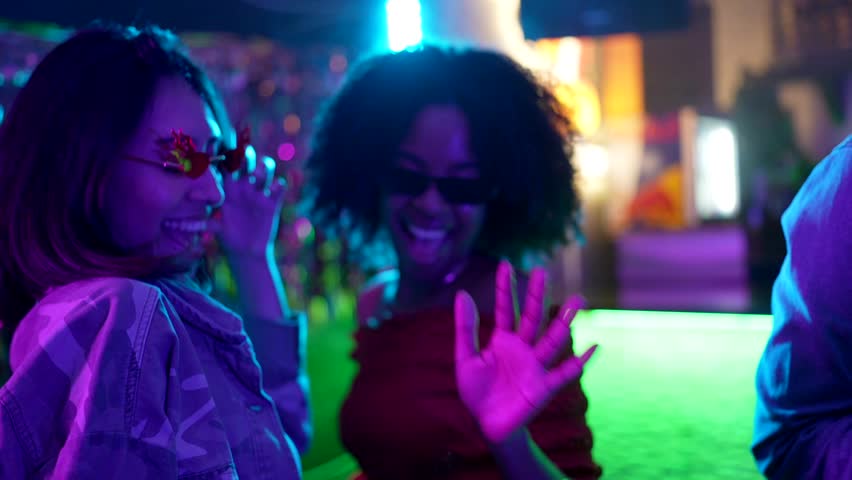 Cheerful diverse female friends with sunglasses smiling and dancing together during a party in a nightclub with vibrant blue and purple neon lights, enjoying the music and lively atmosphere