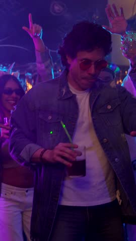 Cool young man in a denim jacket and sunglasses holding a cocktail while dancing with friends at a nightclub party with colorful neon lights, having a great time during a vibrant evening out