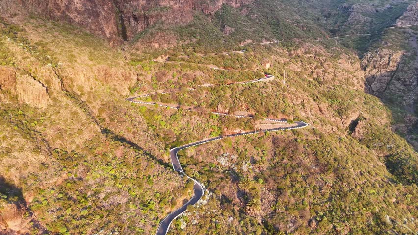 Aerial view of cars navigating a winding mountain road. Autos riding at scenic landscape with lush greenery and rocky terrain under soft sunlight. Automobile journey through beautiful nature route