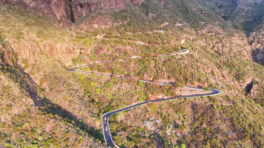 Aerial view of cars navigating a winding mountain road. Autos riding at scenic landscape with lush greenery and rocky terrain under soft sunlight. Automobile journey through beautiful nature route