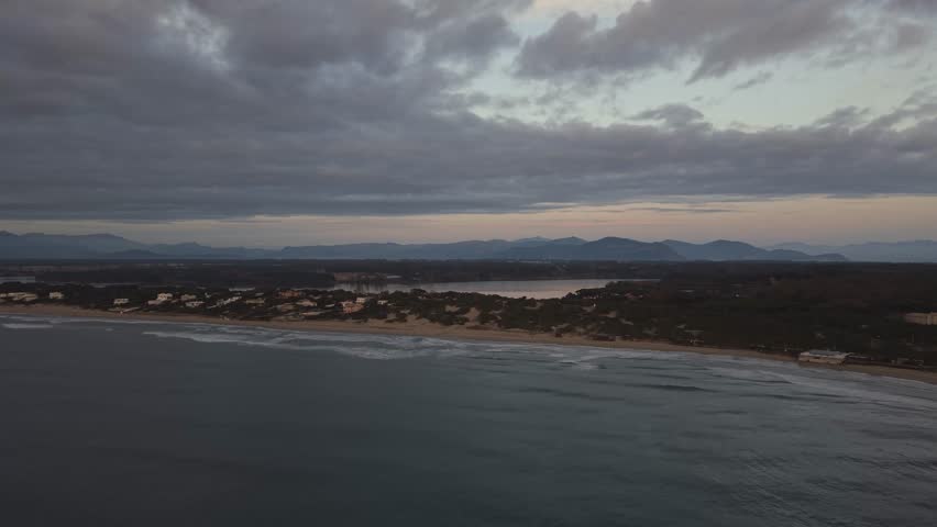 Wide aerial drone view of the coastal strip in Sabaudia, Italy, separating the lake and the Mediterranean Sea. Panoramic perspective of the sandy dunes, lake water, and mountain range under a cloudy sky.