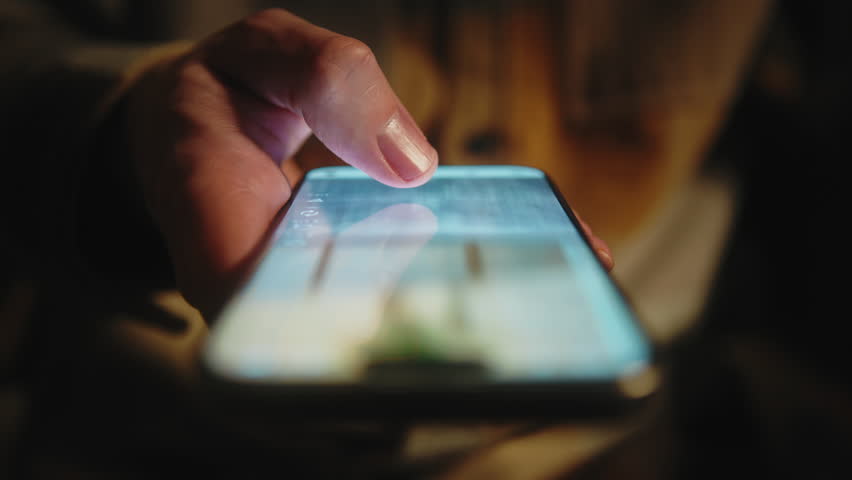 Close-up male hand in casual jacket scrolling social media feed on smartphone at night. Swipe motion blur on phone screen. Evening online browsing, digital lifestyle, content consumption, technology