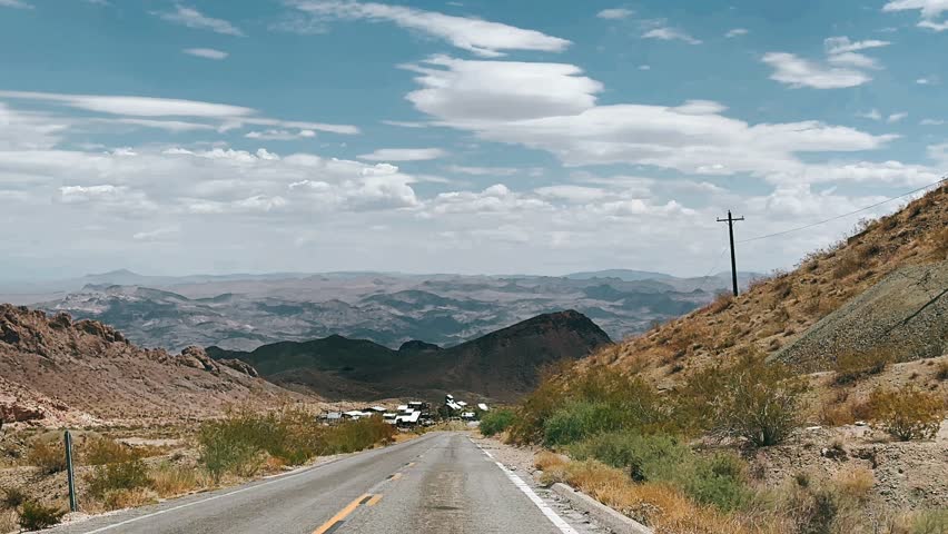 A quiet roadway stretches forward through rugged desert mountains, framed by dry brush, rocky slopes, and layered hills fading into the distance. The asphalt road leads toward a small cluster of buildings nestled between arid ridges, while soft clouds drift across a pale blue sky. The scene captures isolation, scale, and the slow rhythm of travel through an expansive desert landscape shaped by time and terrain.