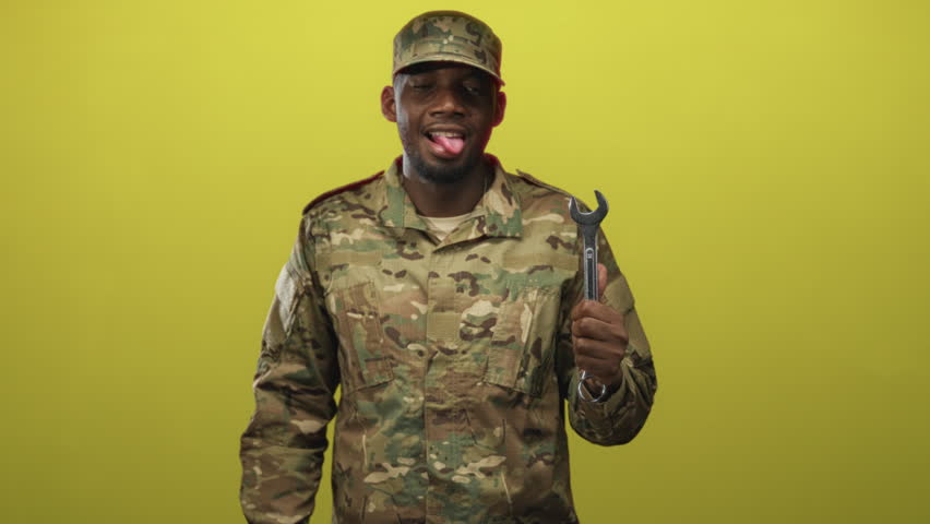 Young black man soldier in camouflage jacket and cap holding large wrench, sticking tongue out in studio; playful pride.