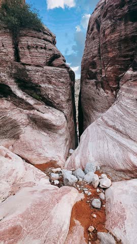 A tight natural corridor cuts between towering sandstone formations, revealing layers of red, cream, and rose-colored rock shaped over time by wind and erosion. The stone walls rise steeply on both sides, their textured striations leading the eye upward toward a bright blue sky dotted with soft clouds. Scattered rocks rest along the canyon floor, emphasizing the raw desert terrain and the dramatic scale of Red Rock Canyon near Las Vegas.