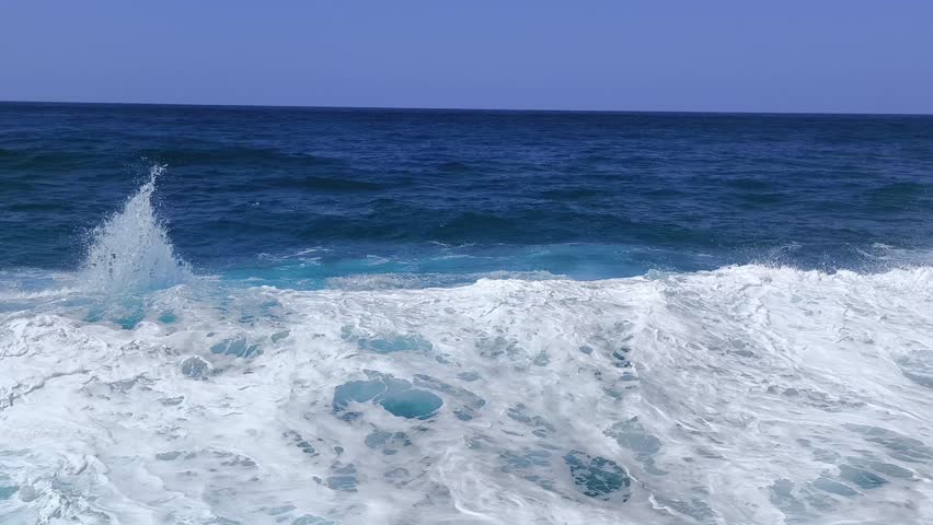 Slow motion ocean waves roll in with bright foam. Deep blue water swirls under a clear sky near the horizon.