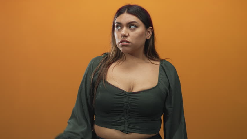 Woman points index finger and shows cleavage in a studio with orange backdrop, wearing green blouse and smirking while posing confidently; confidence.