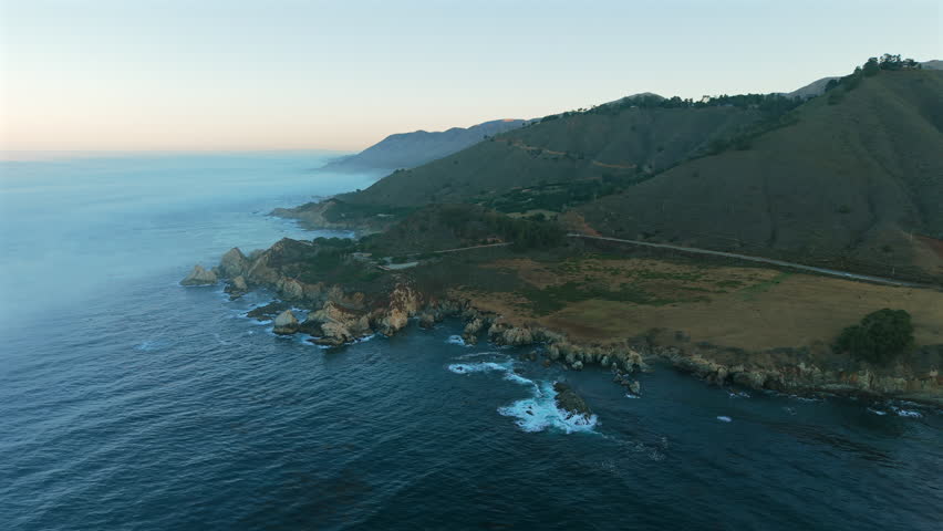 Aerial view of Big Sur California coastline with dramatic cliffs winding coastal highway and deep blue Pacific Ocean captured in soft light creating cinematic landscape scene.