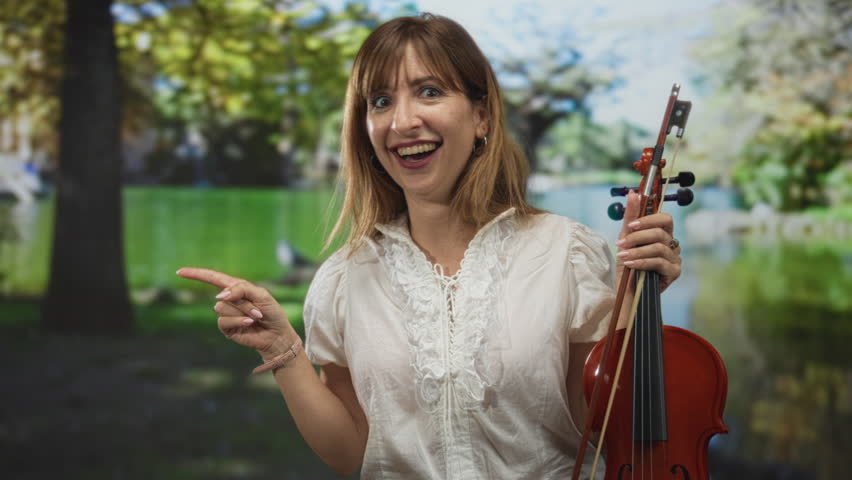 Woman holding violin points index finger in park by a lake while smiling in white blouse; musical joy.