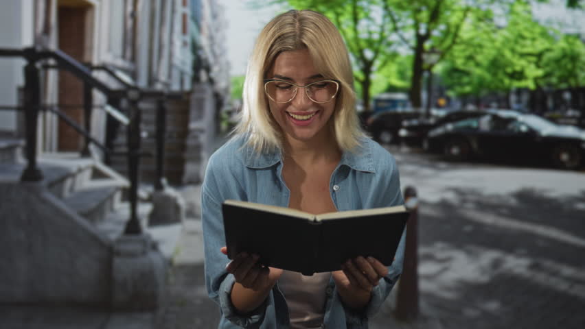 Woman holding open book with both hands, smiling and reading with expressive gestures on a street by building steps and parked cars, wearing glasses and denim shirt; joyful.