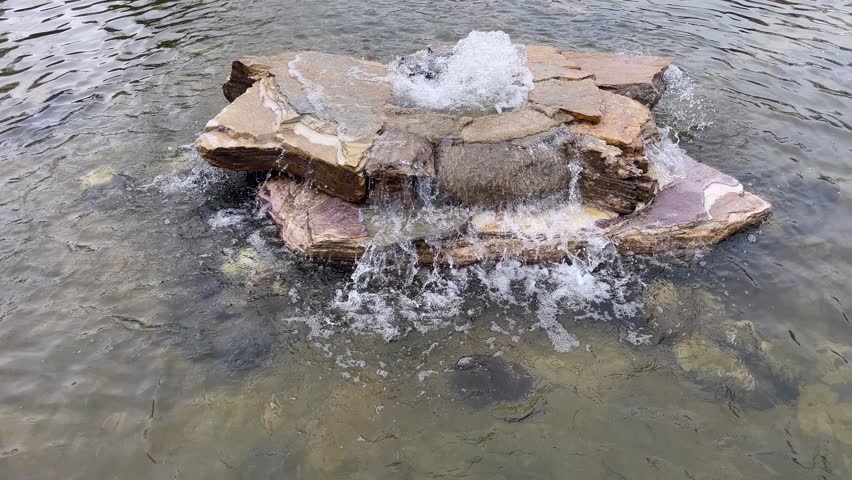 Rock Fountain Natural Pond. A clear, open-air pond with a rock fountain. Ideal for travel and wellness. It showcases a blend of relaxation and landscape design, suitable for themes related to visual appeal, wellness, outdoor recreation, or luxury garden features. A high-angle shot of a tranquil swimming pond with a bubbling rock water feature in the center. Waves spread across the water