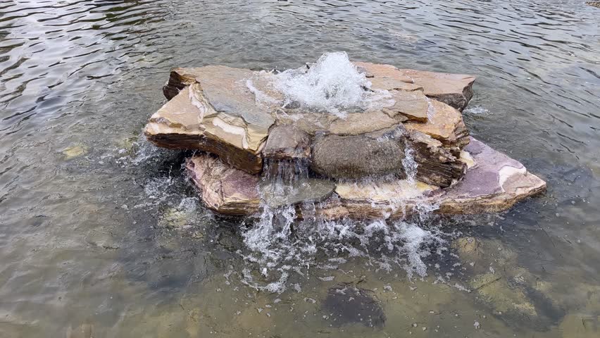 Rock Fountain Natural Pond. A clear, open-air pond with a rock fountain. Ideal for travel and wellness. It showcases a blend of relaxation and landscape design, suitable for themes related to visual appeal, wellness, outdoor recreation, or luxury garden features. A high-angle shot of a tranquil swimming pond with a bubbling rock water feature in the center. Waves spread across the water