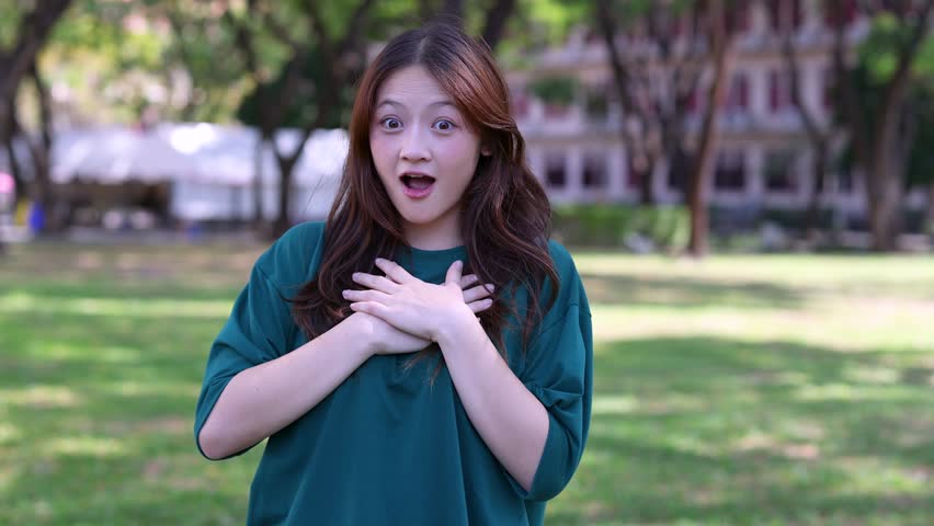 A young Asian woman expresses shock and joy with hands on chest in a park