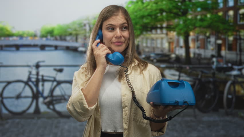 Young blonde woman holds blue telephone handset to ear on cobblestone street by canal bridge in amsterdam city; connection communication urban vibrancy.