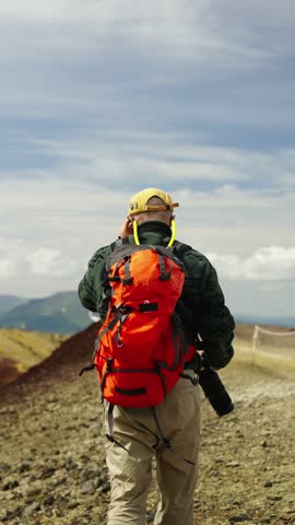 Solo hiker with bright red backpack walking along rocky mountain trail under wide blue sky, capturing scenic wilderness landscape with camera from behind