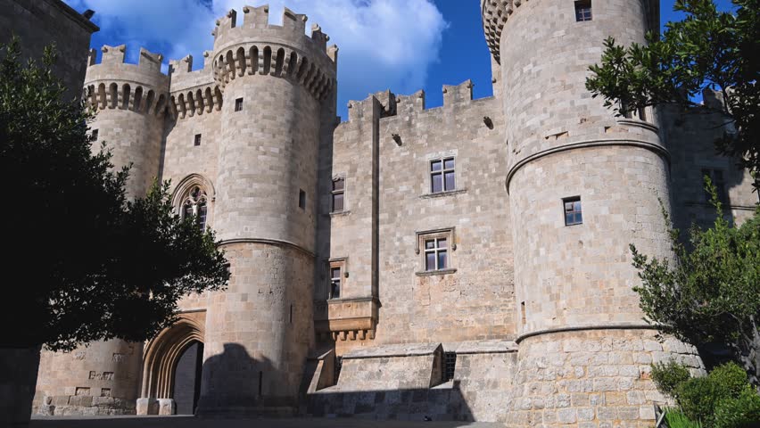 Ground level walking shot moving along the stone paved area near the entrance of the Palace of the Grand Master of the Knights of Rhodes in the town of Rhodes, Greece with fortified walls visible under clear blue sky