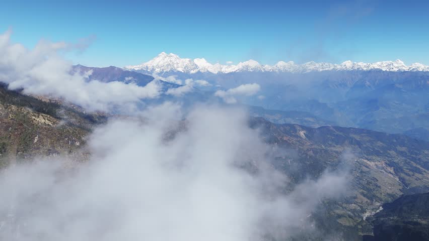 Hanging clouds float above green mountains with Himalaya peaks in the background creating a scenic aerial landscape in Nepal