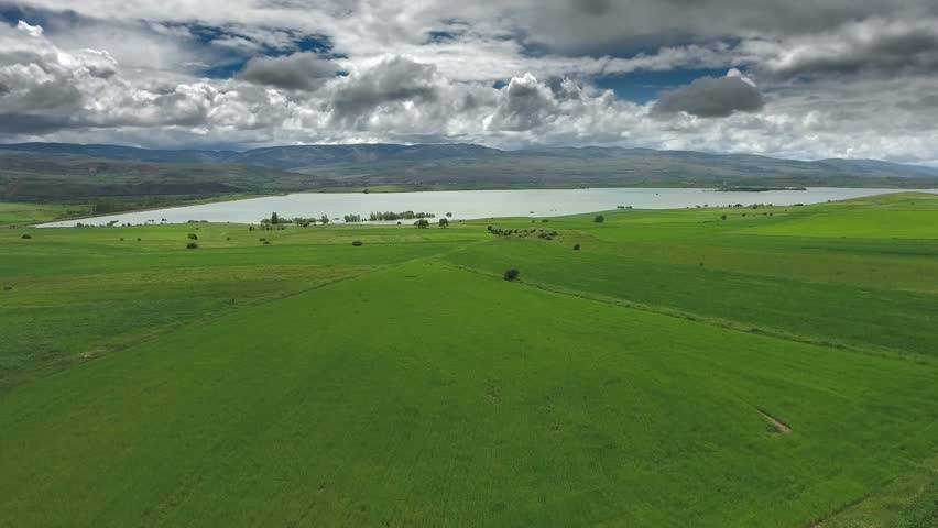 Aerial flight over green cultivated fields heading toward a calm lakeshore under clouds. Wide rural panorama shows patchwork farmland, grassy plains, and distant water basin beneath stormy sky.