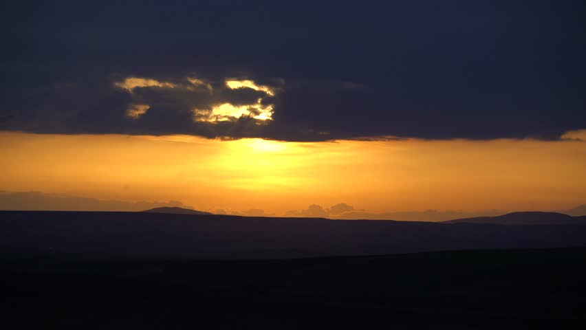 Taklamakan Desert sunset glows beneath heavy cloud bank, golden light over silent dunes. Arid Xinjiang twilight burns through dark canopy, amber sun sinks beyond barren horizon.