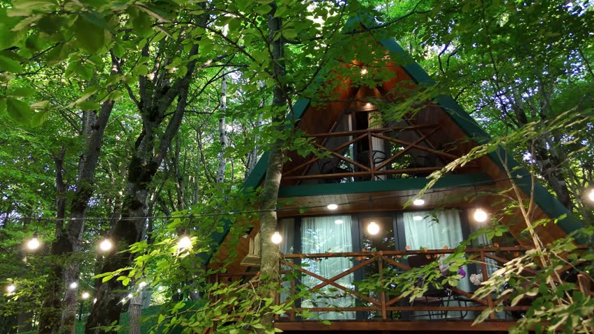 Wooden a frame cabin standing amidst the dense green foliage of a vibrant forest, with warm string lights creating a cozy and magical atmosphere for a perfect glamping escape