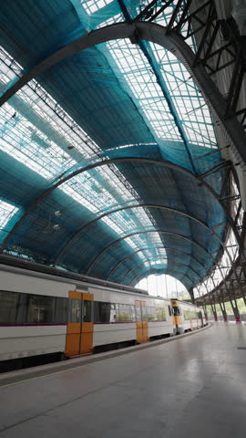 A modern train waits under a large arched roof covered in blue construction netting at a Barcelona station platform.