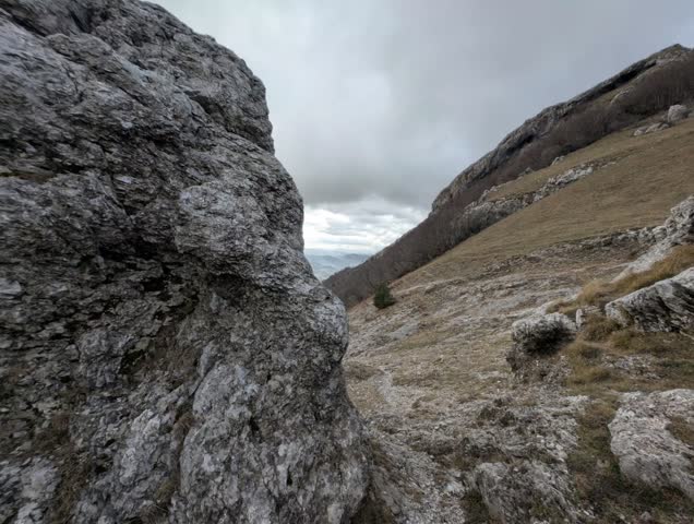 Mountain landscape with large rock formation in the foreground, grassy slopes and hills in the background under a cloudy sky, rugged natural terrain with sparse vegetation