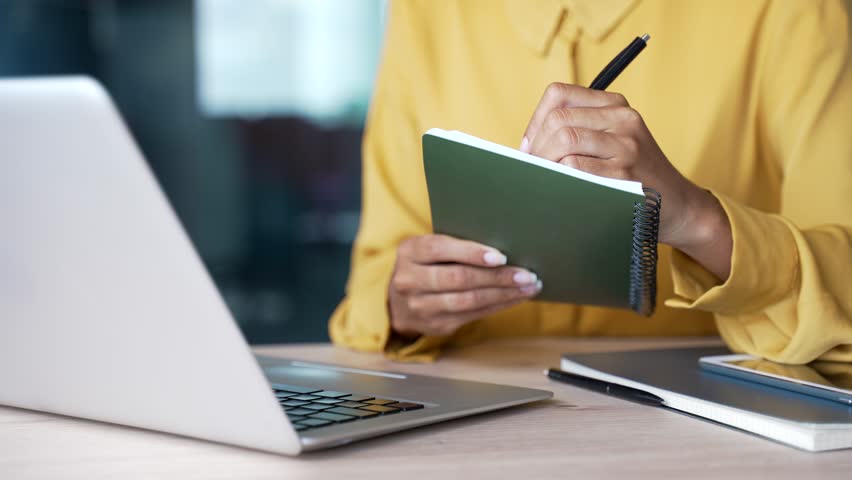 Close-up of businesswoman’s hands writing notes in notebook while using laptop at desk in modern office. Female employee taking notes during remote business meeting, online conference training session