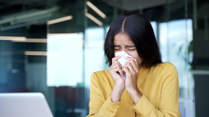 Close-up of sick young businesswoman blowing her nose with a handkerchief in modern office. Unhealthy female employee suffering from cold or flu, coughing and sneezing while feeling tired at work.
