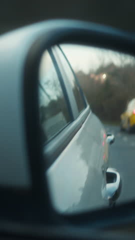 Rear side mirror view of a yellow car following on a curved road through a forested area, suggesting travel or pursuit, vertical video
