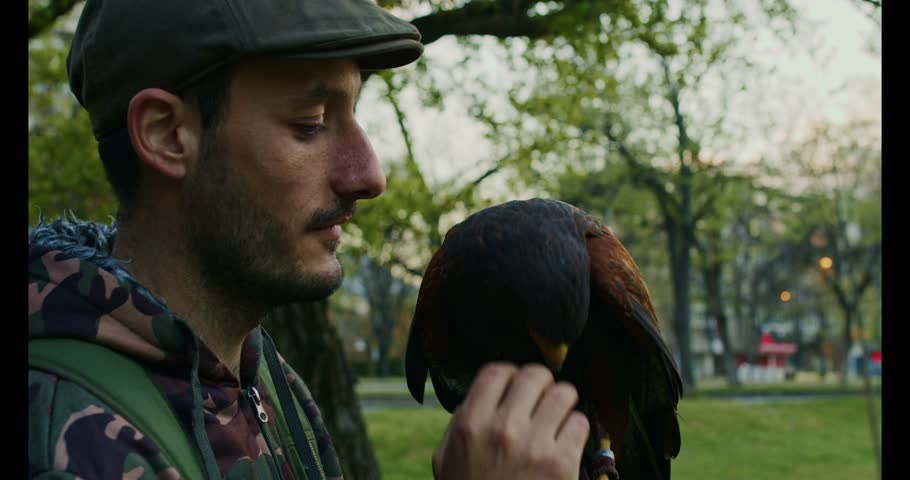 Side profile of a man in cap holding a falcon on his gloved hand outdoors in a leafy park, natural light, shallow depth of field