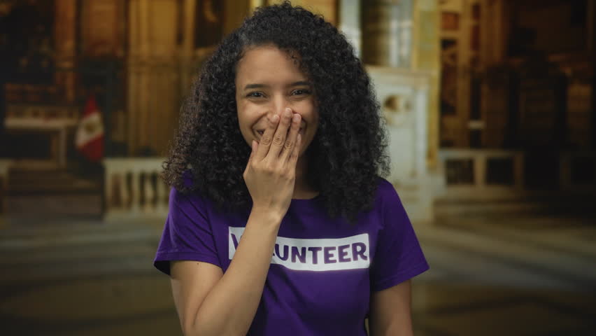 Young latin volunteer woman in a volunteer shirt covers her mouth and laughs inside a church building; joy.