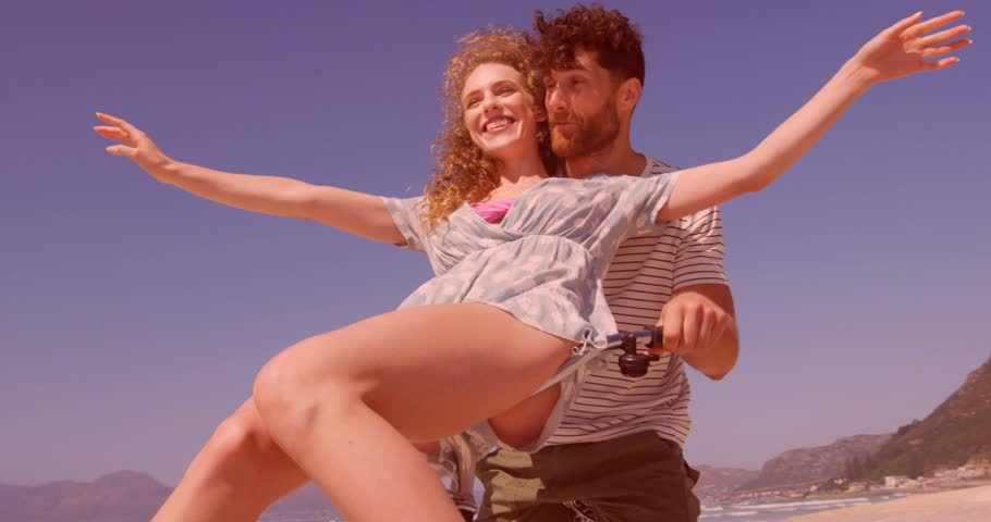 Couple riding bicycle along beach, pedaling motion causing promo hearts rising for romance. Duo, bike, balloons, shoreline, coastal, sunny, playful