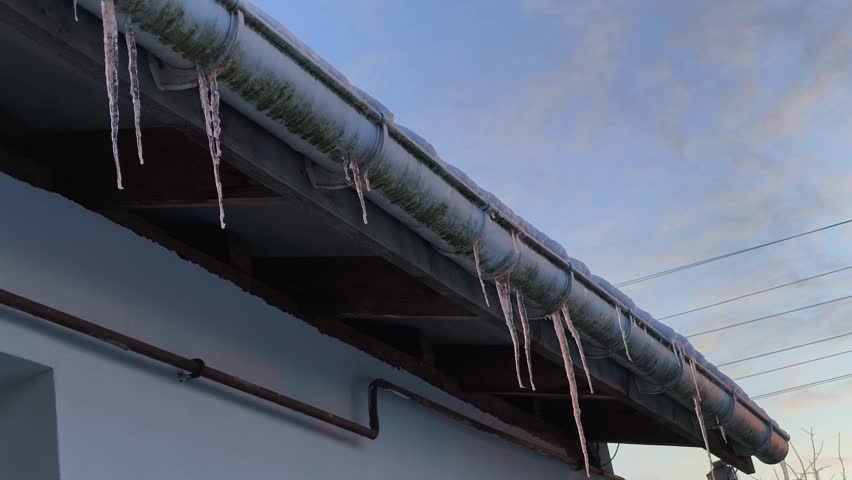 Long icicles dangle from a weathered metal roof gutter, capturing the chill of a winter evening. The scene is set against a serene blue sky with soft clouds, highlighting the beauty of frozen water in
