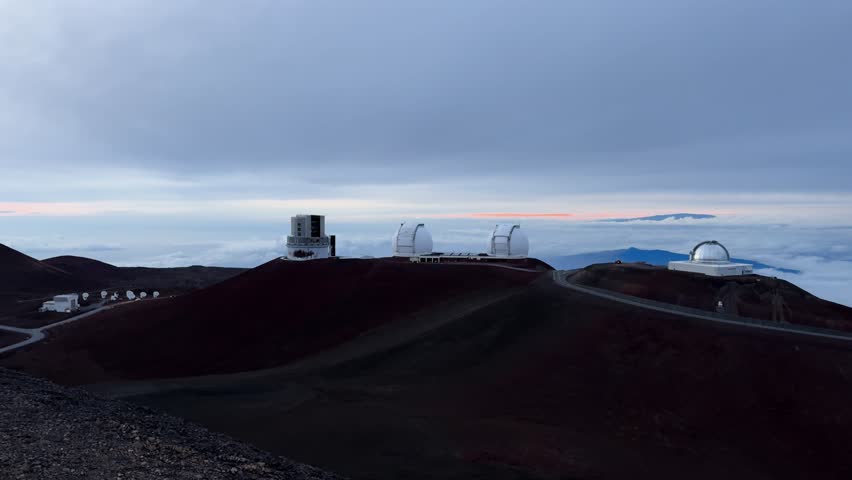View from the Mauna Kea summit in Hawaii, showing multiple observatory domes on volcanic hills, with access roads and cloudy sky.
