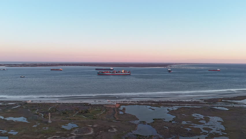 Drone aerial time lapse at sunrise over the east end of Galveston Island showing coastal wetlands, the long rock jetty extending into the Gulf of Mexico, and multiple cargo ships anchored offshore waiting to enter the Houston Ship Channel. Warm morning light reveals tidal pools, marsh patterns, and shipping traffic on the horizon. Strong environmental and maritime themes with clear copy space and layered composition.
