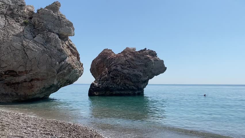 Scenic coastal landscape in Kemer, Turkey featuring impressive rock formations rising from the turquoise Mediterranean Sea. Crystal clear water, pebble beach and bright summer sky create a perfect travel and vacation concept on the Turkish Riviera.
