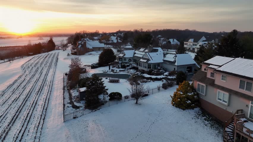 American mansion with frozen pool on cold snowy winter day. Sunset time in rural area of town. Snow-covered roof and agricultural fields at dusk. Aerial view.