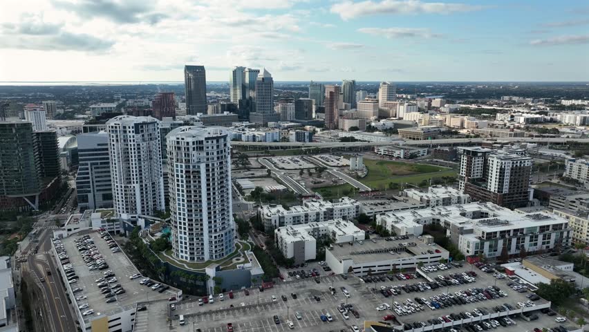 Aerial view of Tampa showing modern residential towers, downtown skyline in distance, highways, parking areas and dense urban development under bright Florida sky. Wide shot.