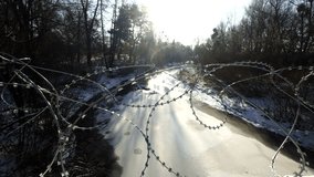 Razor Barbed Wire Coils Over Frozen River In Winter Forest, Sharp Metal Security Fence Protecting Border Area, Military Grade Obstacle Installed Near Snowy Water Stream, Defensive Perimeter With Concertina Wire In Natural Wilderness, Safety Barrier Blocking Access During Cold Weather Season, Protective Boundary Under Bright Sunlight In Winter Landscape, Steel Razor Wire Protection Over Ice Covered River, Security Measures In Scenic Snowy Woodland Environment, Sharp Edged Metal Fencing For Area Defense, Dangerous Obstruction On Ice Surface In Nature, Protective Installation Preventing Passage Through Frozen Riverway, Border Security Fence In Wild Winter Environment - Powered by Shutterstock - Get 15% off with code: PIKWIZARD15
