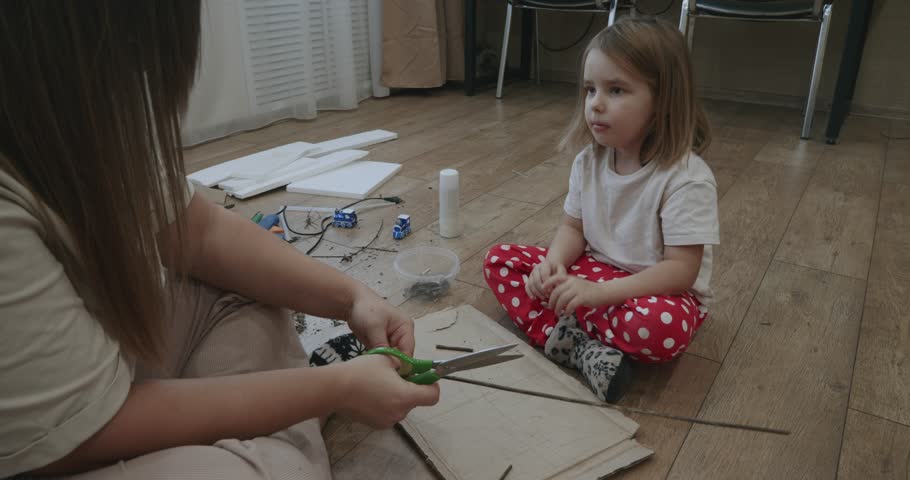 Young girl sitting on the floor watching her mother cut materials with scissors for a homemade project. Loving family spending quality time together creating arts and crafts at home.