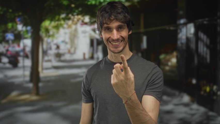 Man displays cheeky grin and shows middle finger in a shaded urban street lined with trees; defiance.