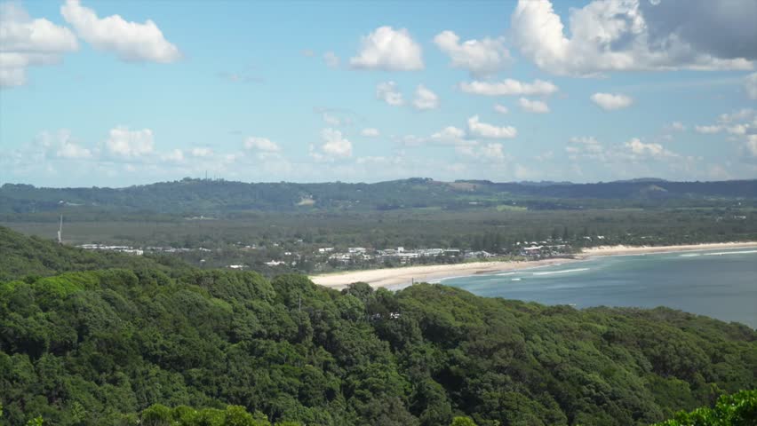 Wide coastal view looking north from the Byron Bay headland, showing forested hills, town outskirts and a sandy beach curving along the shoreline, Byron Bay, Australia.