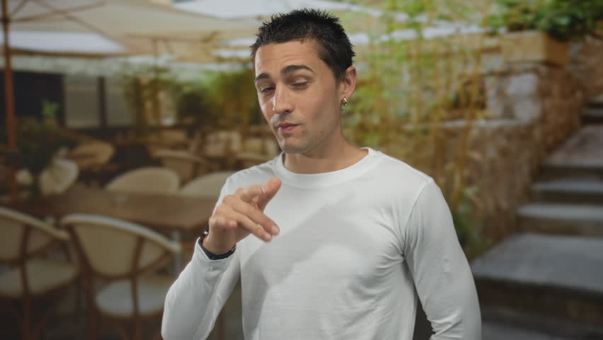 Young hispanic man in white shirt gesturing confidently on a restaurant terrace with greenery in the background, enjoying an outdoor coffee ambiance.