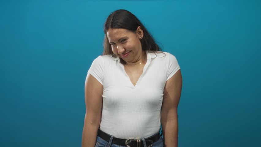 Woman in white shirt raising hands and smiling in a studio with teal blue backdrop; playful confident ease.