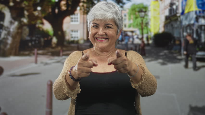 Woman points finger to camera on street, smiling with visible finger and bracelets, wearing cardigan and black tanktop; playful encouragement.