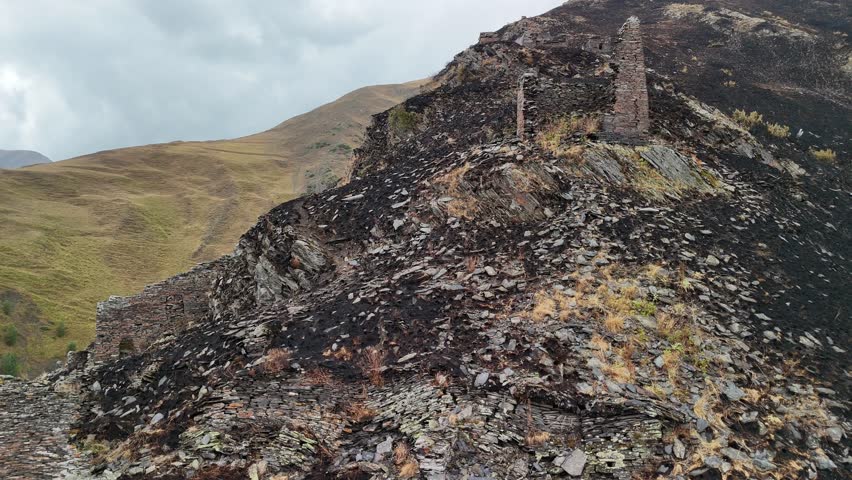 Aerial perspective revealing the aftermath of a fire on a rocky mountain slope, where the ancient stone ruins of a watchtower stand amidst the scorched, blackened earth under an overcast sky