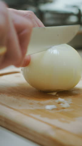 Close-up of hands beginning to slice a white onion on a wooden board in a kitchen, preparing fresh ingredients for cooking, vertical video