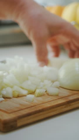Close-up of hands finely chopping a white onion on a wooden board in a bright kitchen, preparing ingredients for cooking, vertical video