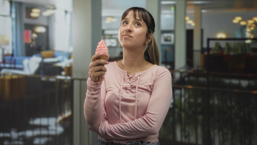 Young woman holding a pink icecream cone in a building, arm crossed while gazing upward and smiling; playful joy.